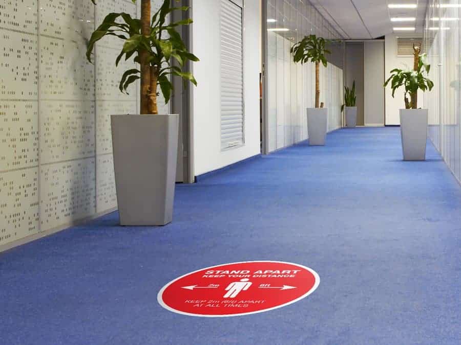 Social Distancing Sticker for Carpet Tiles that says "Stand Apart Keep Your Distance" in an office hallway