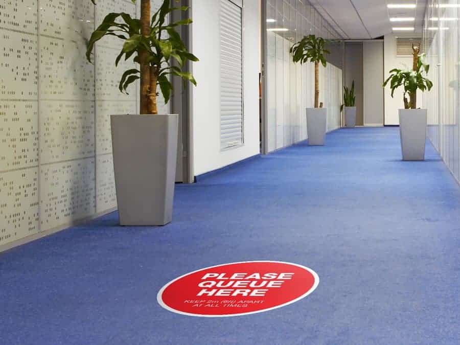 Social Distancing Sticker for Carpet Tiles that says "Please Queue Here" in an office hallway