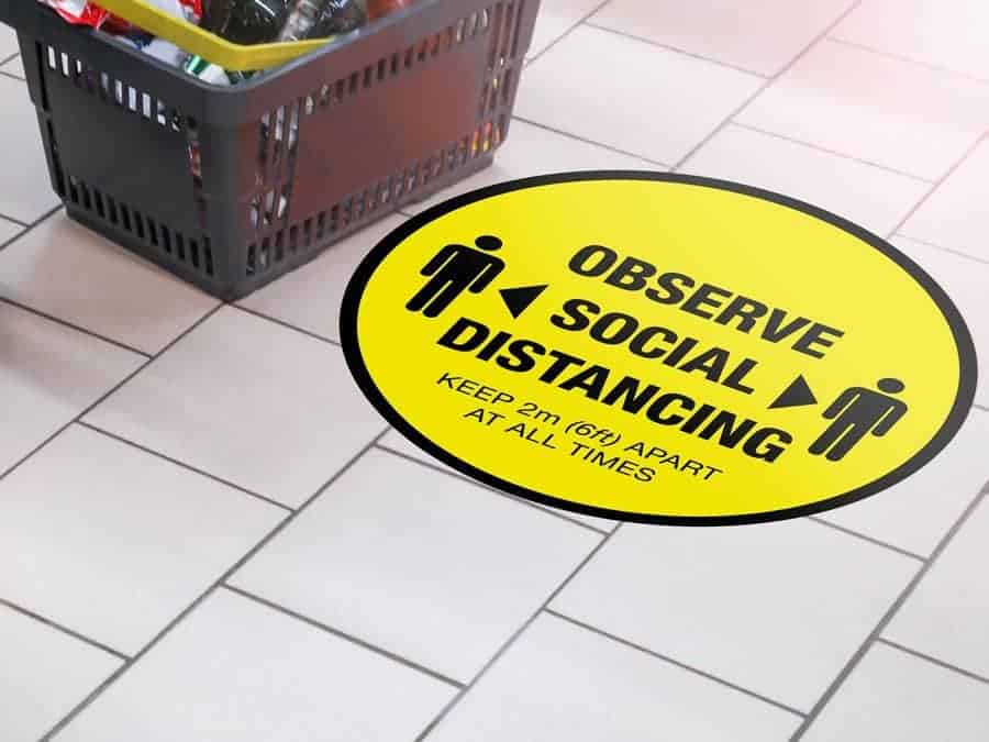 Round yellow Social Distancing Sticker that says "Observe Social Distancing" on tiles next to a shopping basket