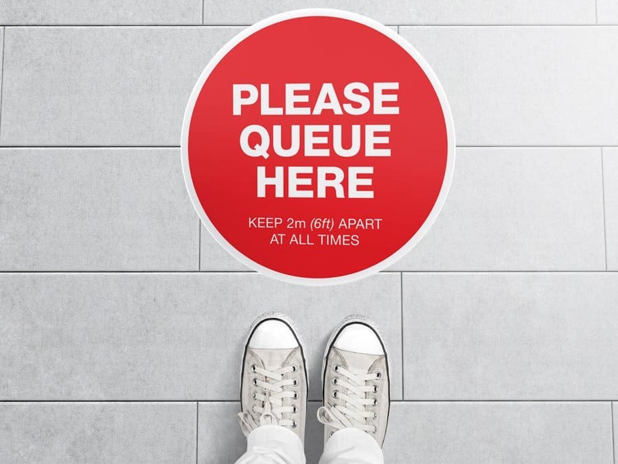 Round red Social Distancing Sticker that says "Please Queue here" on tiles