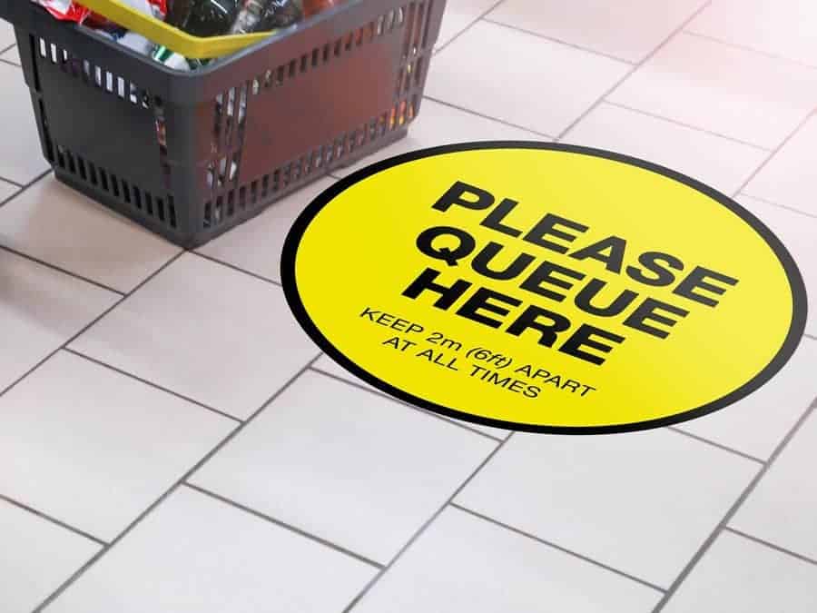 Round yellow Social Distancing Sticker that says "Please Queue here" on tiles next to a shopping basket