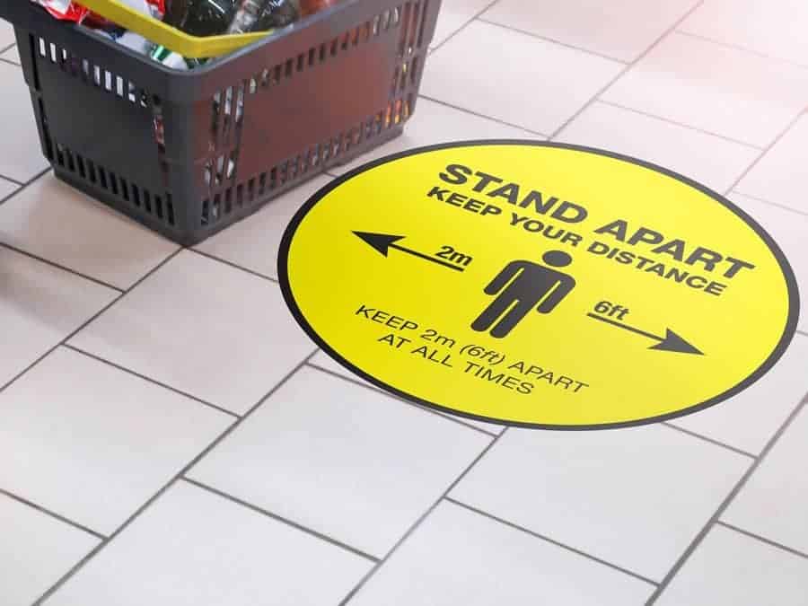 Round yellow Social Distancing Sticker that says "Stand apart keep your distance" on tiles next to a shopping basket