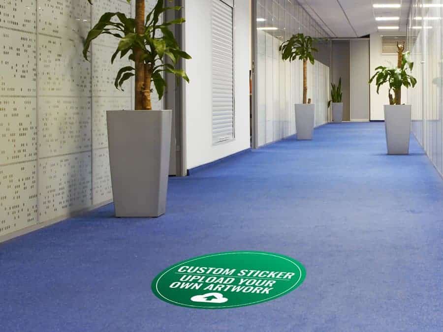 A round green and white social distancing sticker on a carpeted office hallway, with the words "custom sticker - upload your own artwork"