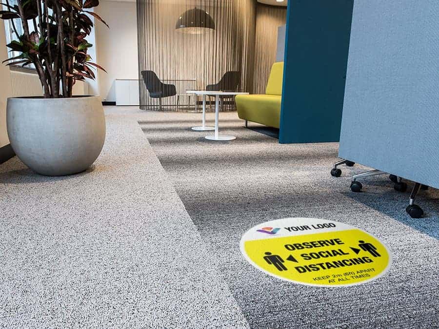 yellow and black social distancing sticker for carpet branded with a logo with the words "Please queue here", in an office hallway