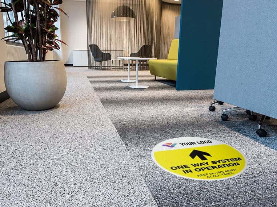 yellow and black social distancing sticker for carpet branded with a logo with the words "one way system in operation", in an office hallway