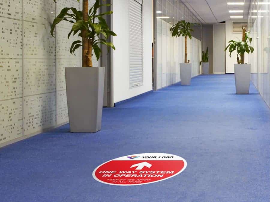 Red and white social distancing sticker for carpets branded with a logo that reads "one way system in operation", inside an office hallway