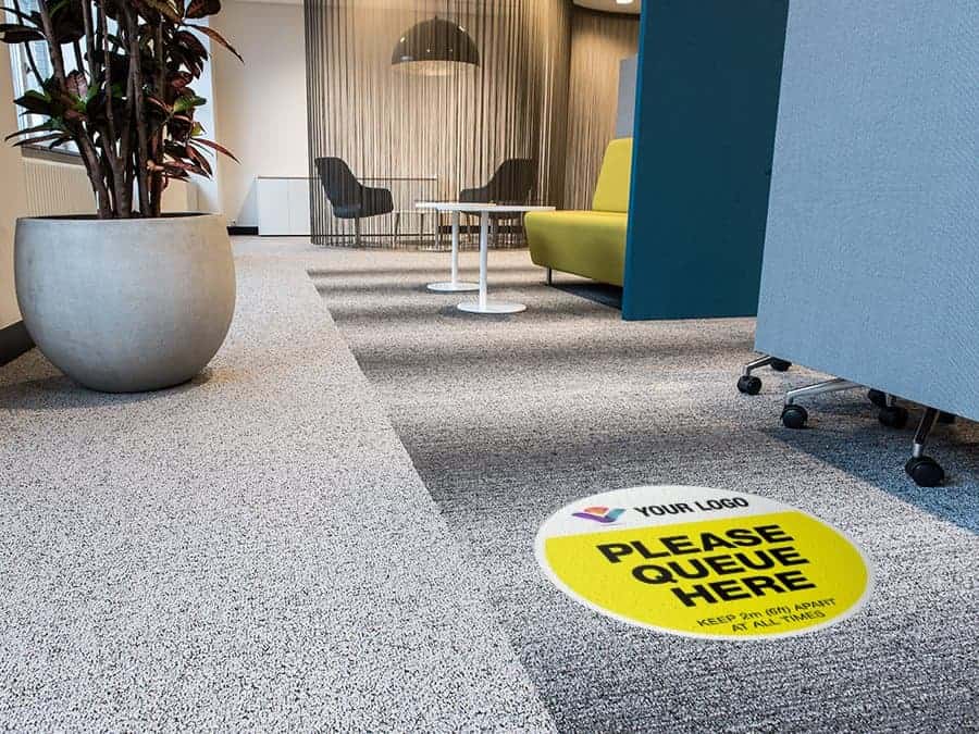 yellow and black social distancing sticker for carpet branded with a logo with the words "Please queue here", in an office hallway