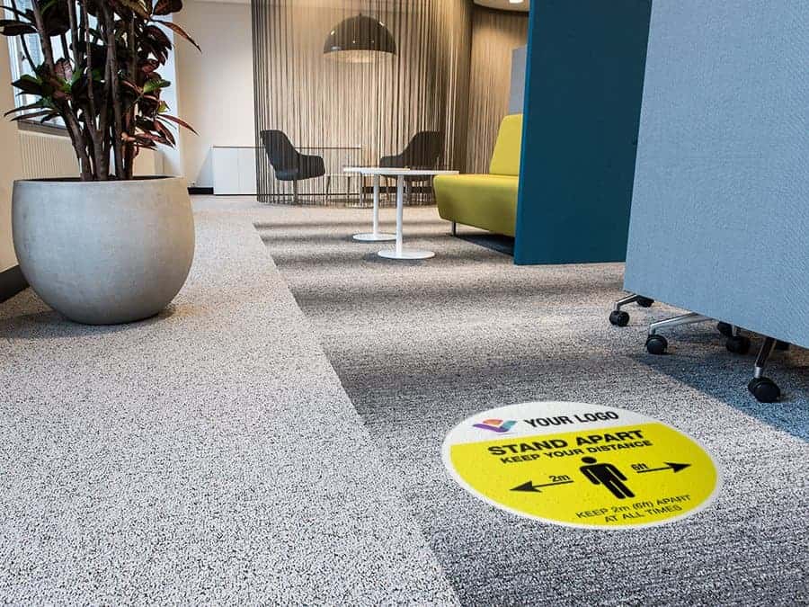 yellow and black social distancing sticker for carpet branded with a logo with the words "Please queue here", in an office hallway