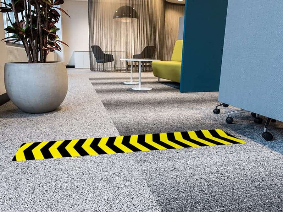 A long, rectangular and yellow and black social distancing sticker with chevrons, on a carpeted office floor