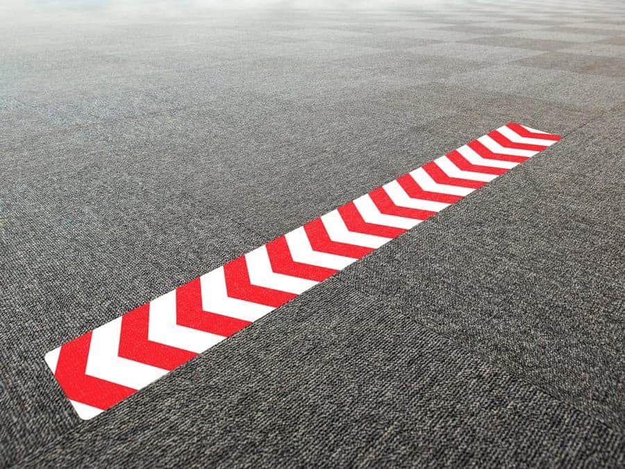 A long, rectangular and red and white social distancing sticker with chevrons, on office carpet tiles