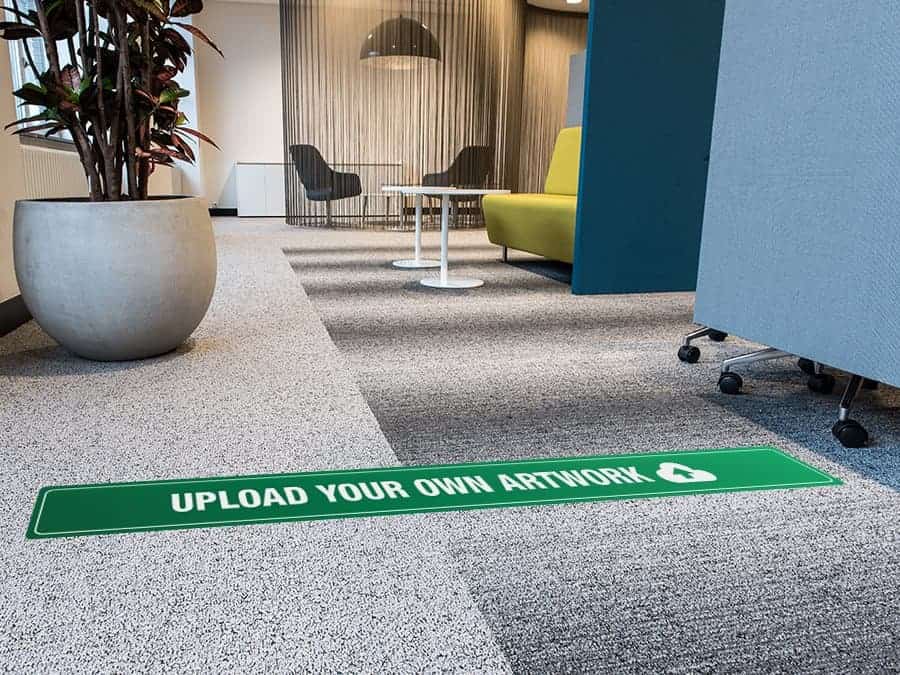 A long, rectangular and green and white social distancing sticker that says "upload your own artwork", on a carpeted office floor