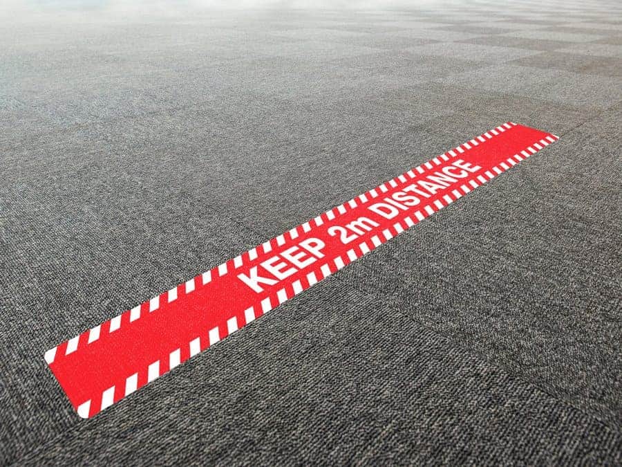 A long, rectangular and red and white social distancing sticker with the words "keep 2m distance", on office carpet tiles