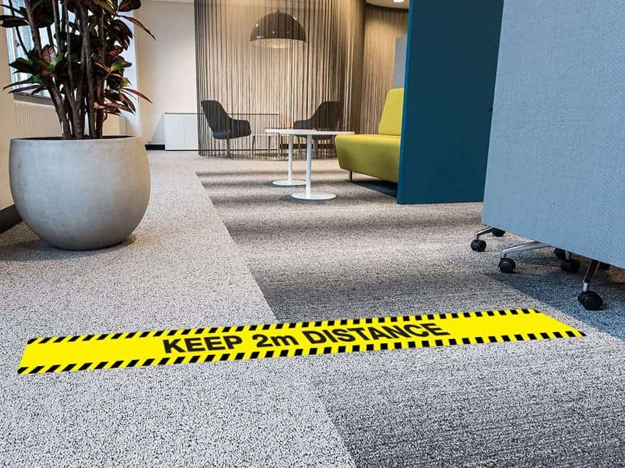 A long, rectangular and yellow and black social distancing sticker with the words "Keep 2m Distance", on a carpeted office floor