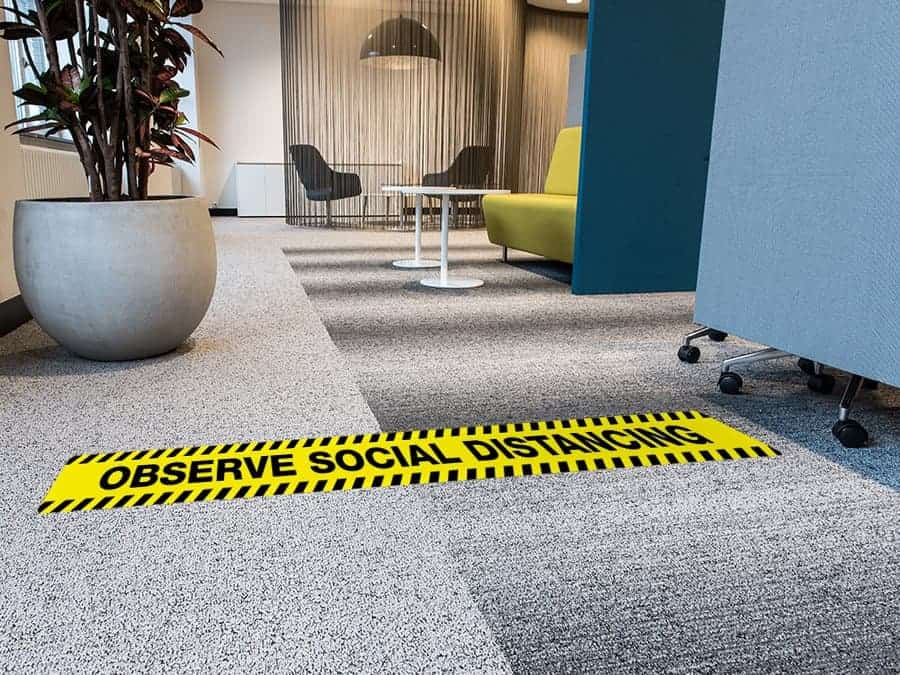 A long, rectangular and yellow and black social distancing sticker with the words "Observe Social Distancing", on a carpeted office floor