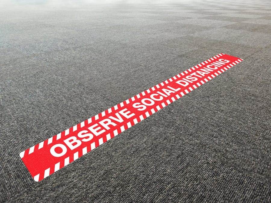 A long, rectangular and red and white social distancing sticker with the words "Observe Social Distancing", on office carpet tiles