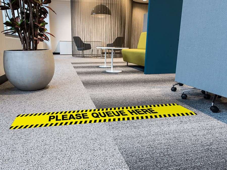 A long, rectangular and yellow and black social distancing sticker with the words "please queue here", on a carpeted office floor