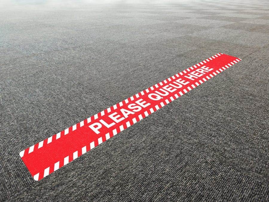 A long, rectangular and red and white social distancing sticker with the words "Please Queue Here", on office carpet tiles