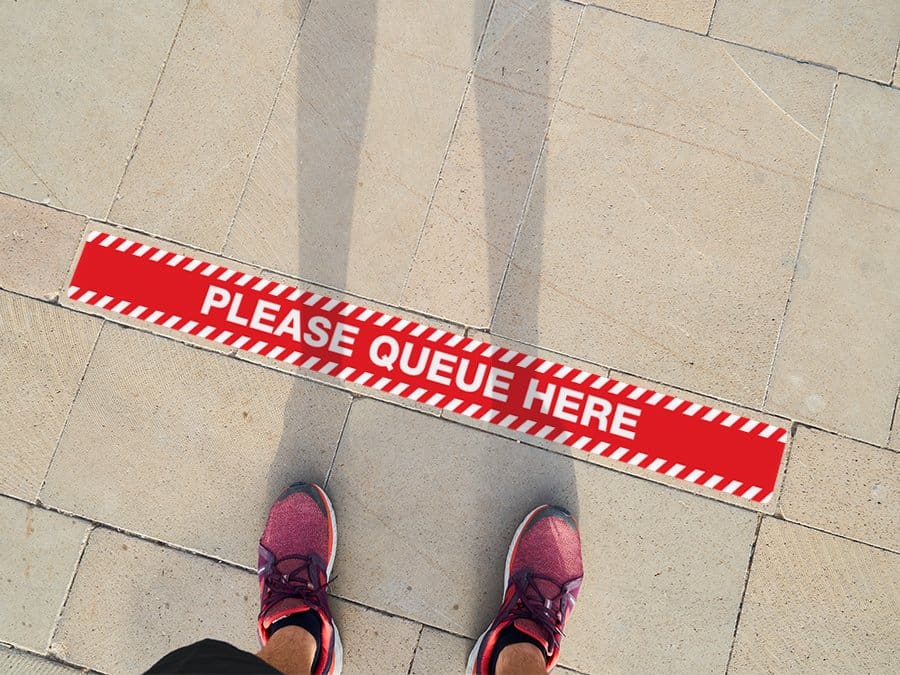 Man standing in front of a social distancing sticker on a pavement with the words "Please Queue Here"